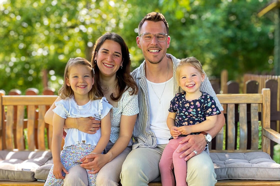 Eine lächelnde vierköpfige Familie sitzt draußen auf einer Holzbank und genießt gemeinsam einen sonnigen Tag.