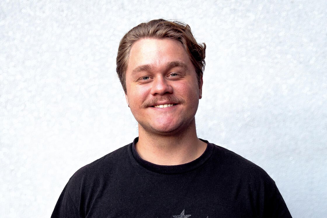 Ein lächelnder Mann mit kurzen braunen Haaren und Schnurrbart, der ein schwarzes T-Shirt trägt und vor einem hell strukturierten Hintergrund steht.