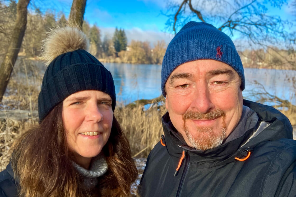 Ein Mann und eine Frau mit Wintermützen, die im Freien an einem See lächeln, im Hintergrund sind Bäume und ein blauer Himmel zu sehen.