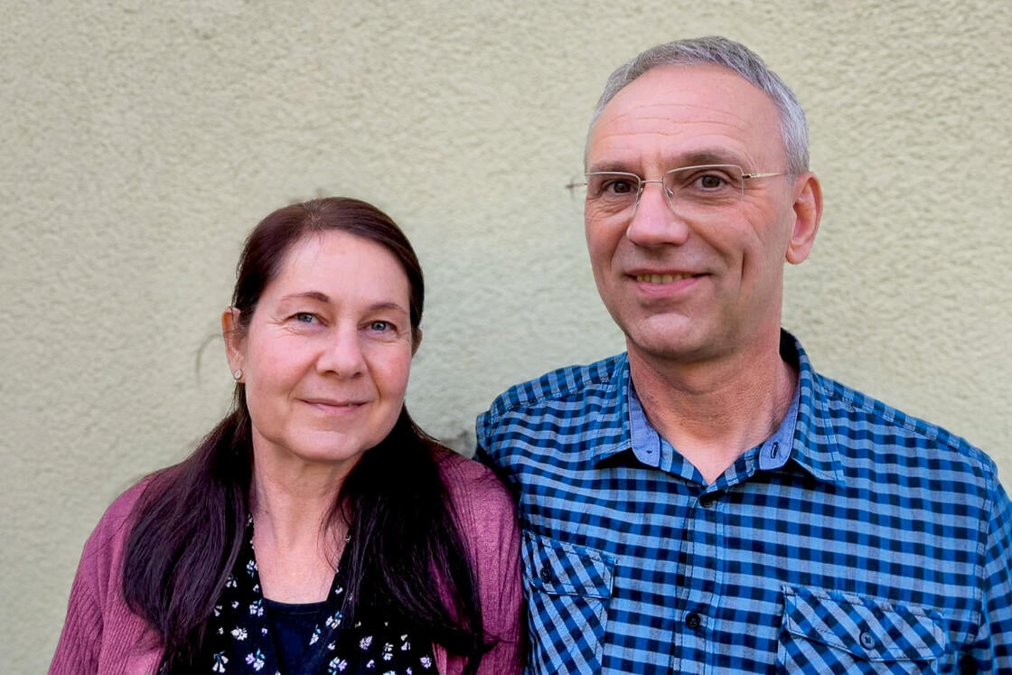 Eine Frau und ein Mann, die lächelnd dicht beieinander vor einer hellen, strukturierten Wand stehen.
