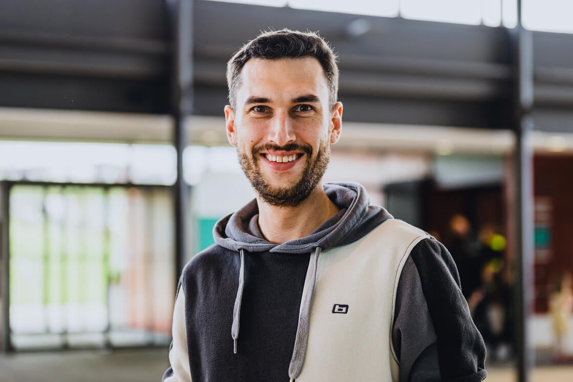 Ein lächelnder Mann mit kurzen dunklen Haaren und Bart, der einen zweifarbigen Kapuzenpulli trägt, steht in einem Innenraum vor einem unscharfen Hintergrund.
