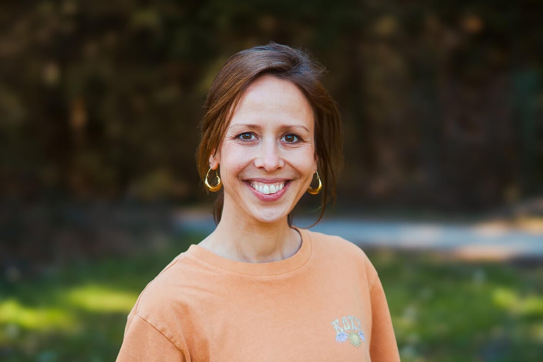 Eine Frau mit braunen Haaren in einem orangefarbenen Shirt, die im Freien lächelt, im Hintergrund ist verschwommenes Grün zu sehen.