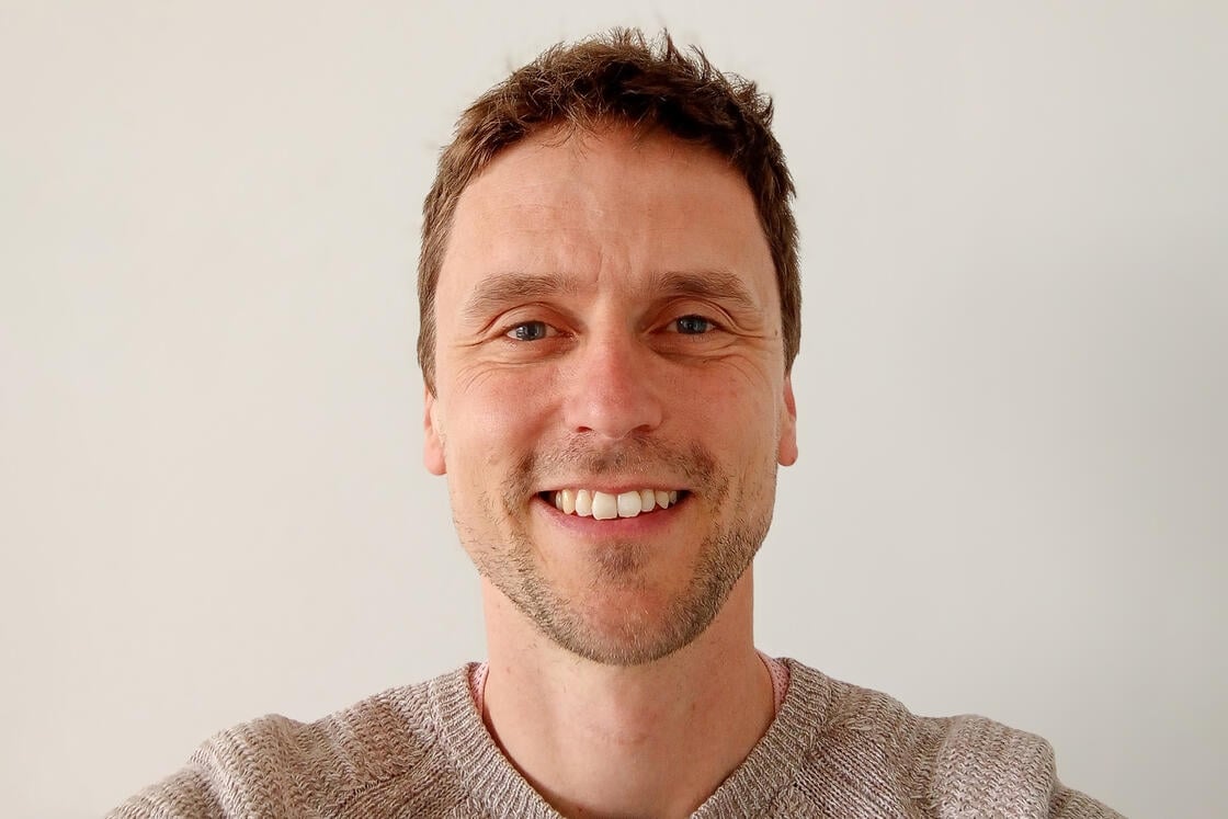 Ein Mann mit kurzen braunen Haaren lächelt vor einem schlichten weißen Hintergrund in die Kamera.