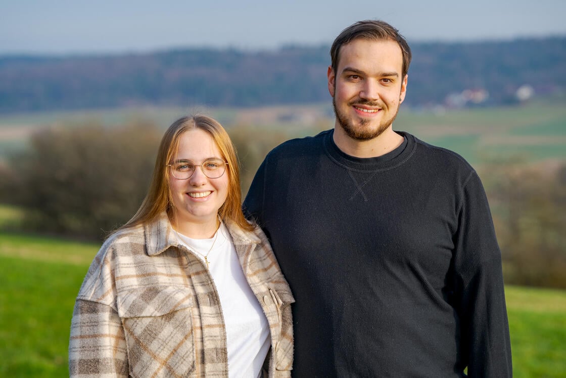 Zwei Menschen stehen gemeinsam im Freien, lächeln, im Hintergrund sind eine grüne Wiese und Bäume zu sehen.
