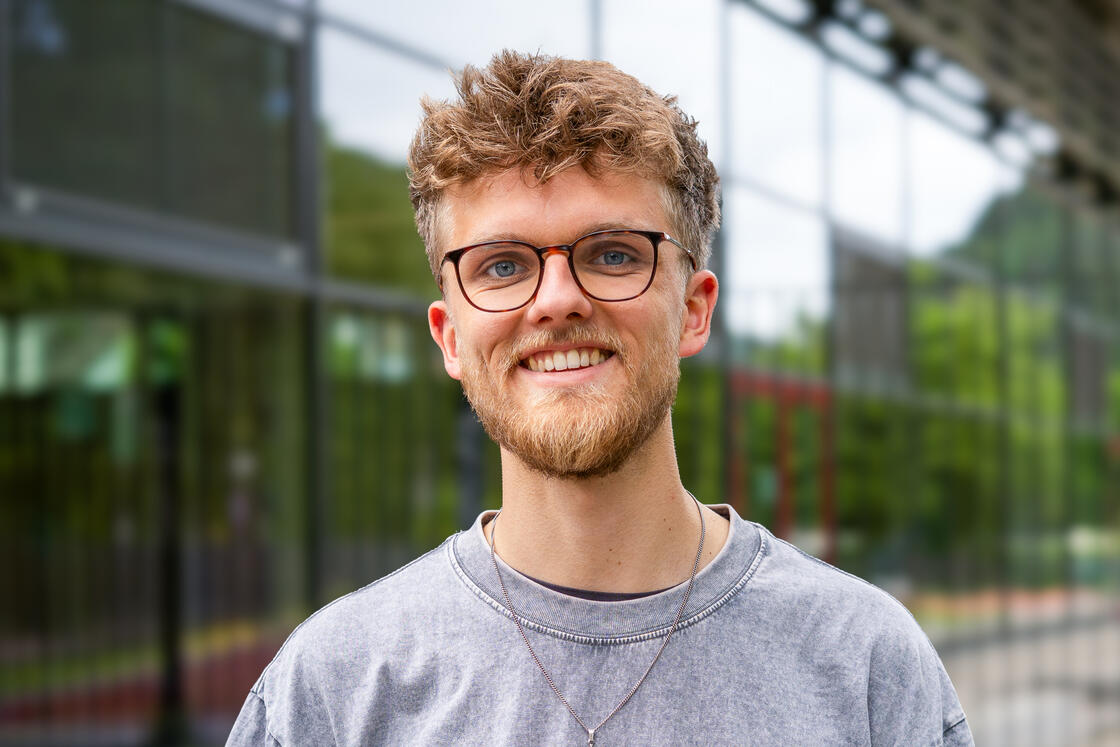 Ein lächelnder junger Mann mit Brille und Bart, der draußen vor einem Glasgebäude steht.
