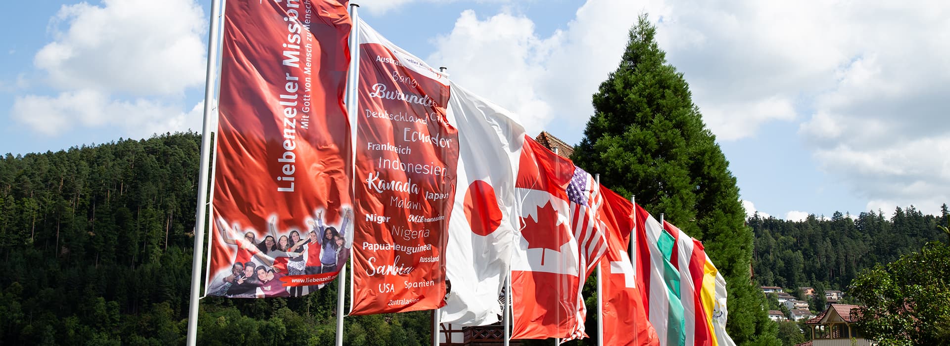 Eine Reihe internationaler Flaggen, darunter die von Kanada und Japan, die im Freien vor dem Hintergrund von Bäumen und Häusern gehisst sind.