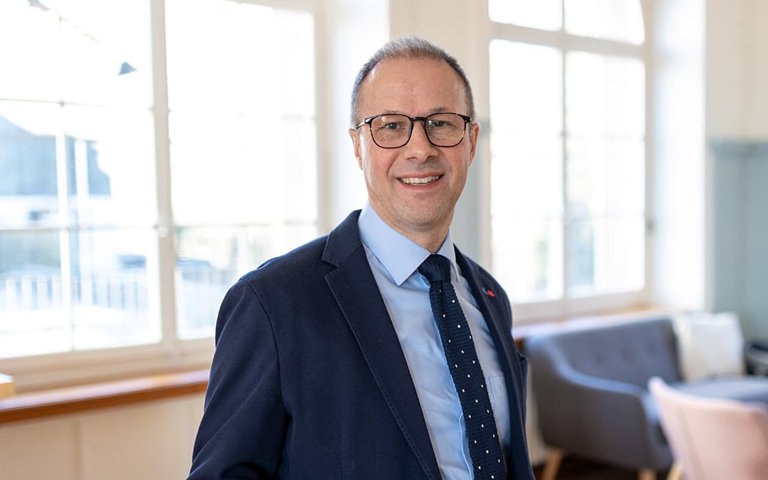 Ein Mann im Anzug und mit Brille lächelt in einem hellen Raum mit großen Fenstern und modernen Möbeln.
