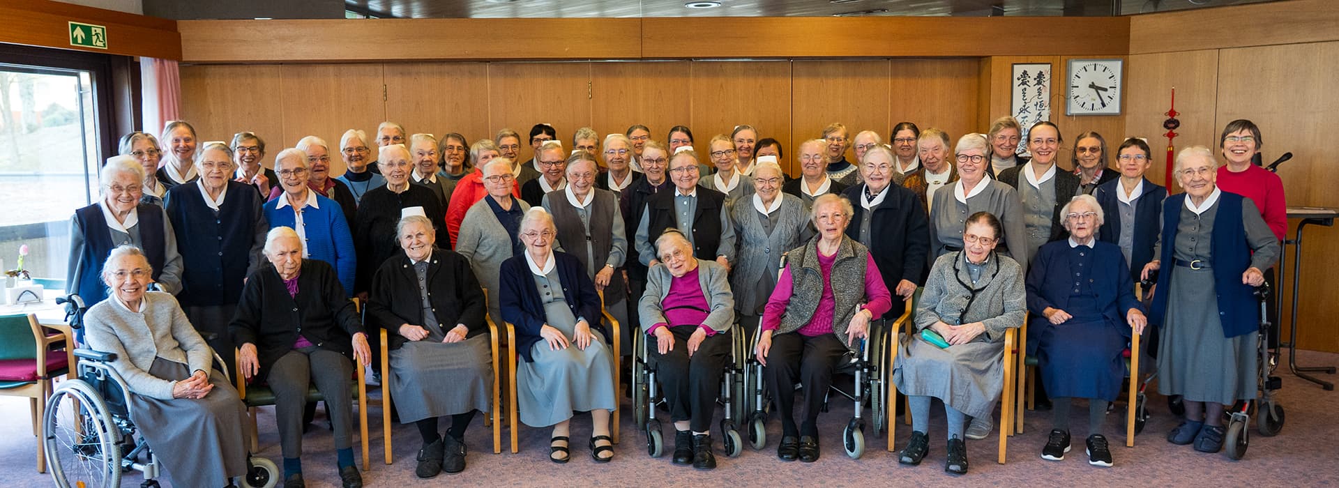 Eine Gruppe älterer Frauen, von denen einige im Rollstuhl sitzen, posiert gemeinsam und lächelt in einem Gemeinschaftsraum mit Holzwänden.