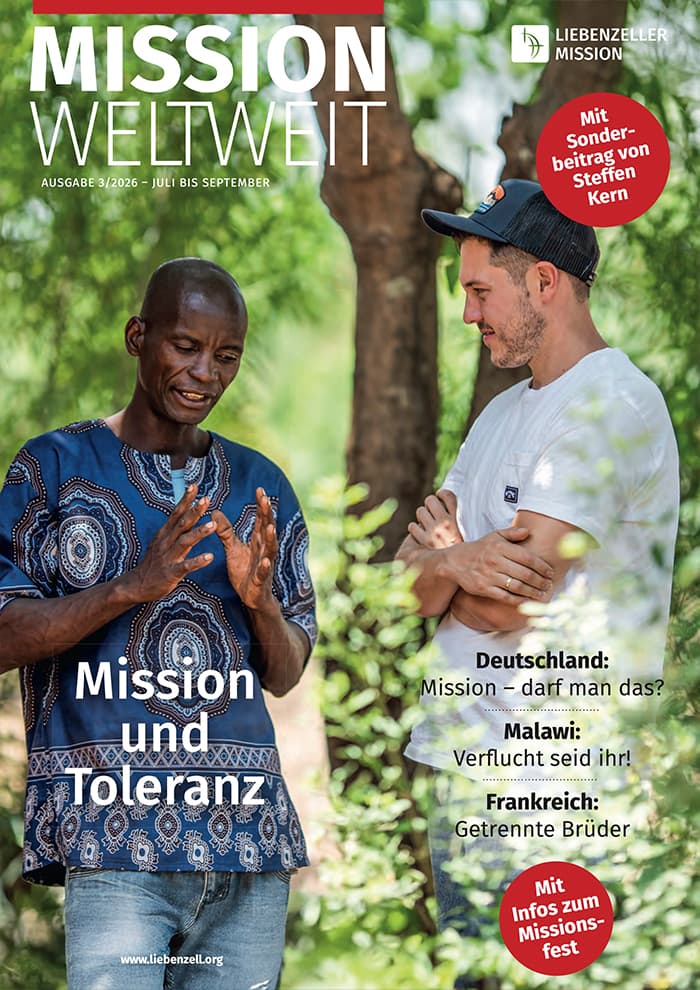Zwei Männer stehen im Freien und unterhalten sich; einer von ihnen gestikuliert beim Sprechen, während im Hintergrund ein Zeitschriftentext über Mission und Toleranz eingeblendet wird.