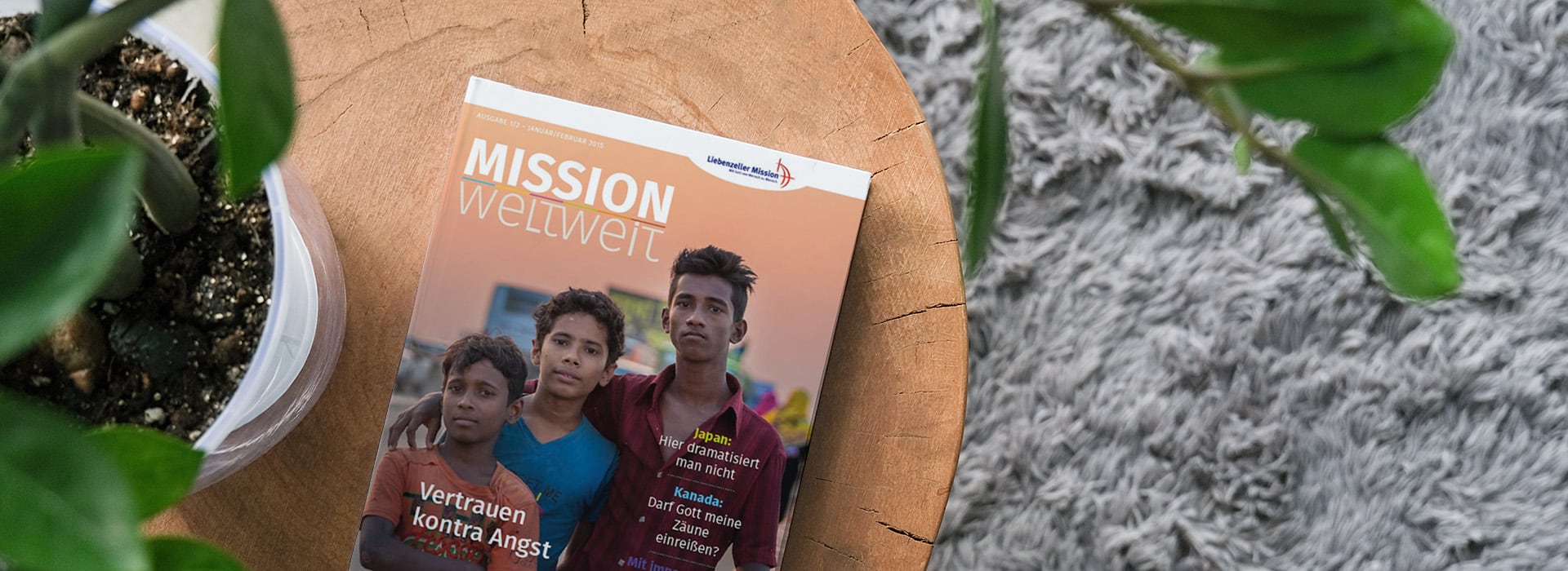 Eine deutsche Zeitschrift mit dem Titel „Mission Weltweit“, auf deren Titelseite drei Jungen zu sehen sind; sie liegt auf einem Holztisch neben einer Pflanze.