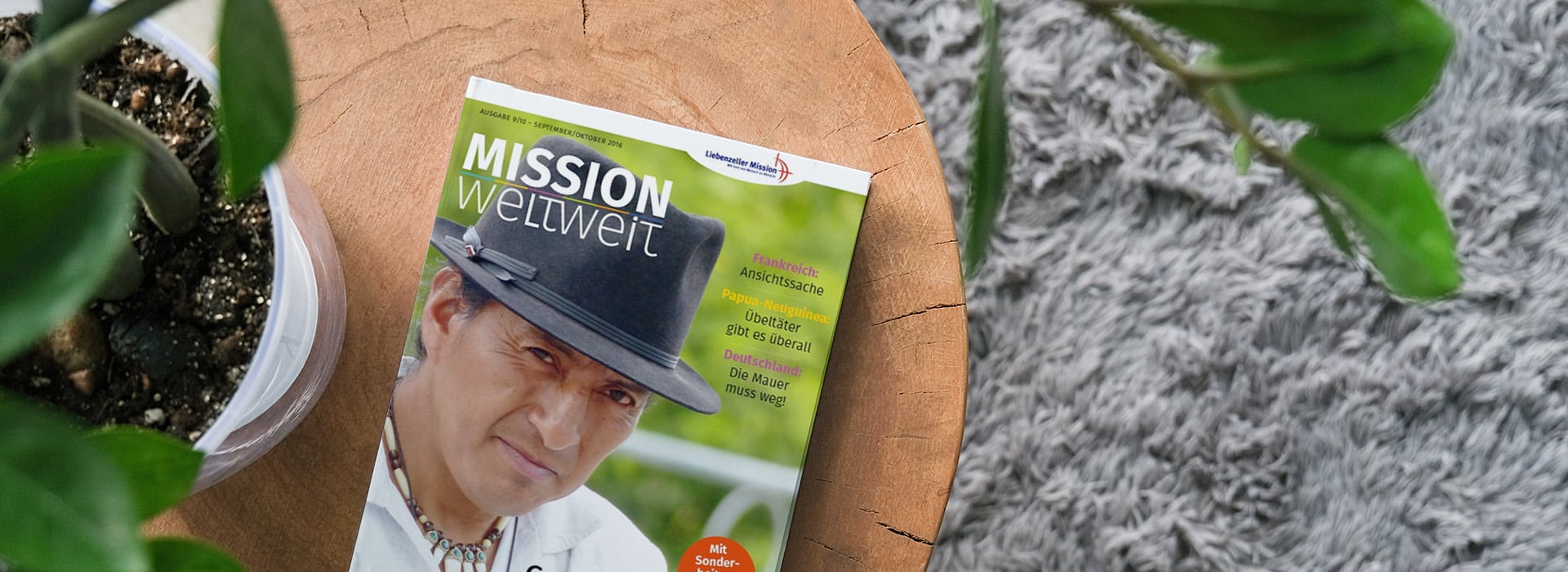 Zeitschrift mit dem Titel „Mission Weltweit“, auf deren Titelbild ein Mann mit Hut auf einem Holztisch zu sehen ist, neben einer Pflanze und einem grauen Teppich.