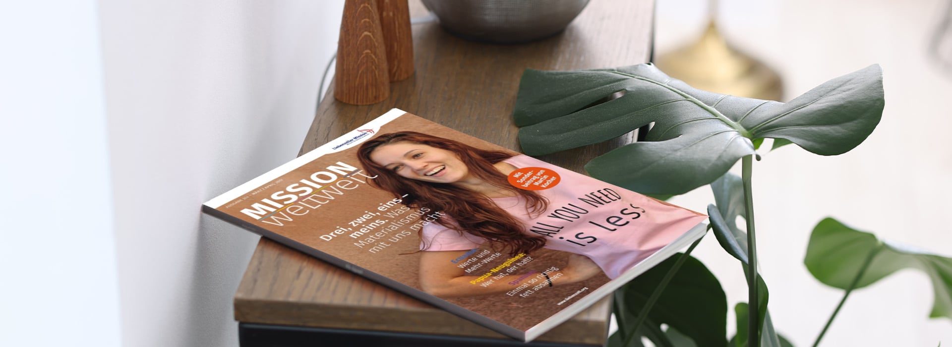 Eine Zeitschrift mit dem Titel „MISSION twenty“, auf deren Titelseite eine lächelnde Frau zu sehen ist, liegt auf einem Holztisch neben einer grünen Pflanze.
