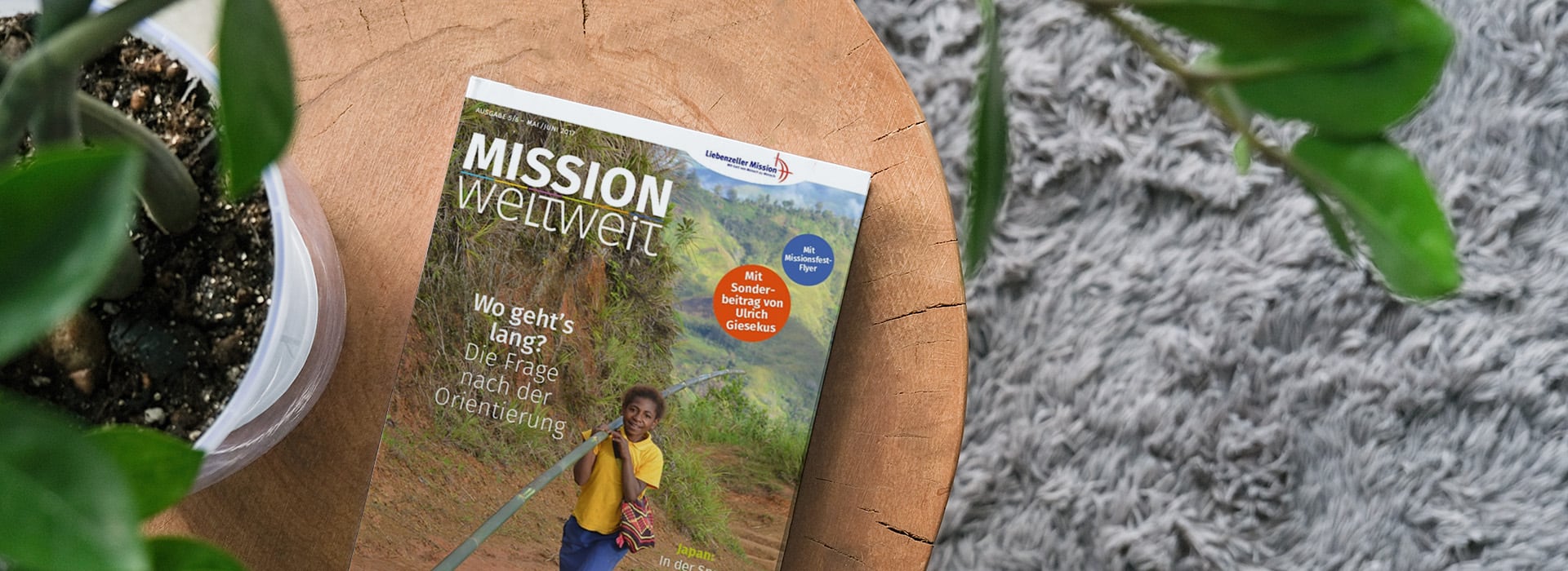 Auf einem Holztisch neben einer Topfpflanze und einem grauen Teppich liegt eine deutsche Zeitschrift mit dem Titel „Mission weltweit“.