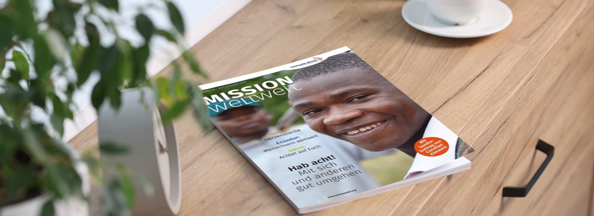 Eine Zeitschrift mit dem Titel „MISSION weltweit“, auf deren Titelseite ein lächelnder Mann zu sehen ist, liegt auf einem Holztisch neben einer Pflanze und einer Kaffeetasse.