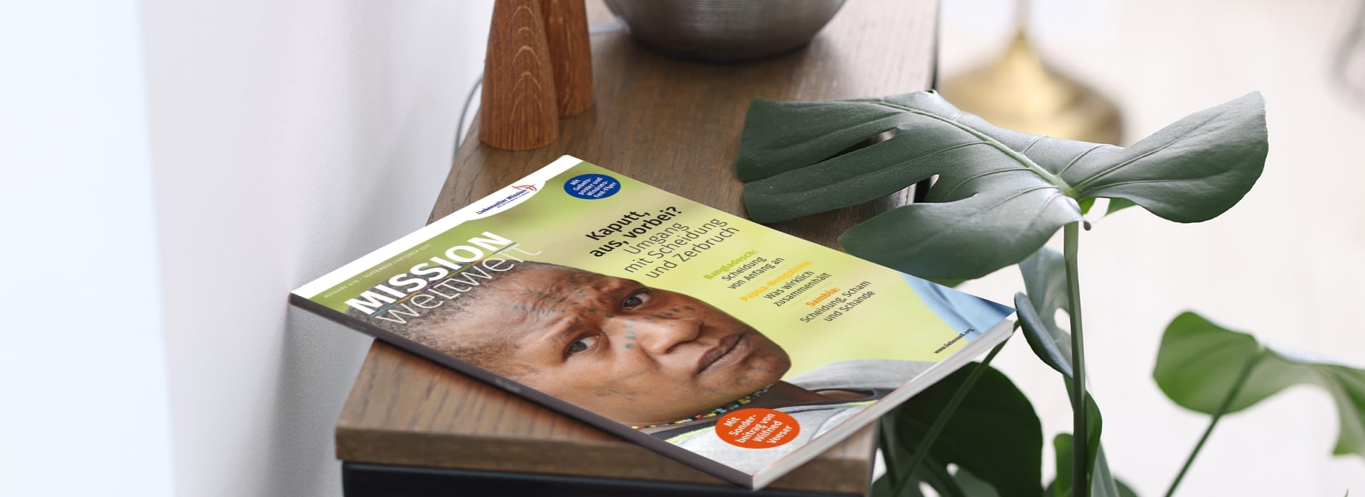 Die Zeitschrift „MISSION weltweit“ mit einer Nahaufnahme eines Gesichts, die auf einem Holzregal neben einer grünen Pflanze liegt.