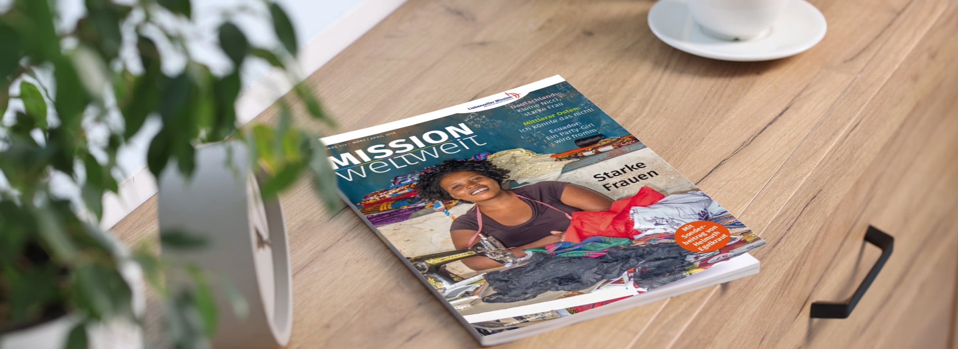 Auf einem Holztisch neben einer Pflanze und einer Tasse liegt eine Zeitschrift mit dem Titel „MISSION weltweit“, auf deren Titelseite eine lächelnde Frau zu sehen ist.