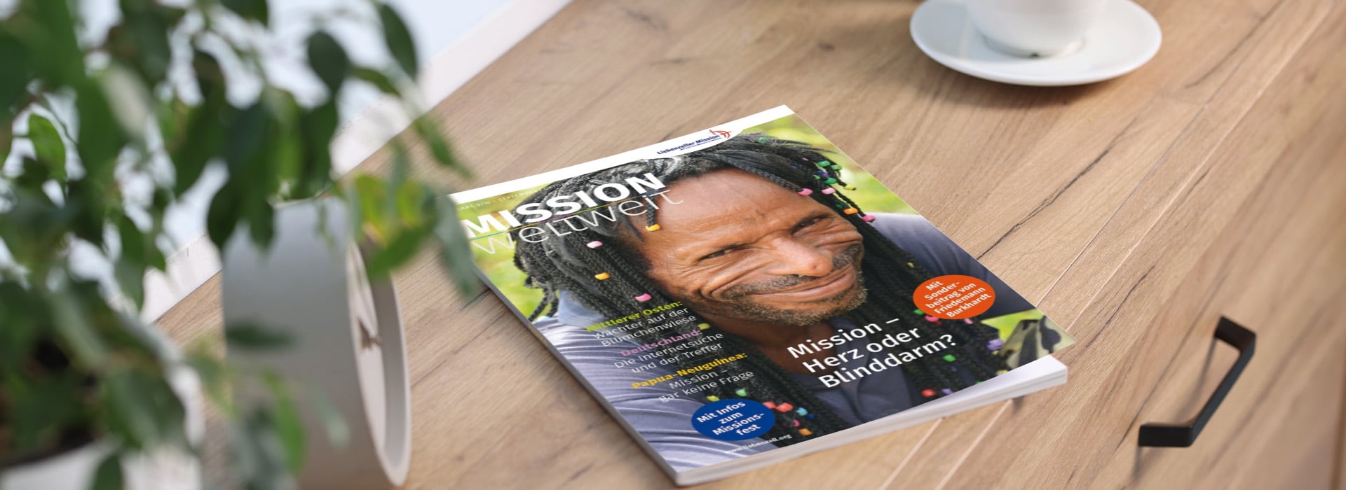 Auf einem Holztisch neben einer Pflanze und einer weißen Tasse liegt eine Zeitschrift, auf deren Titelseite eine lächelnde Person zu sehen ist.