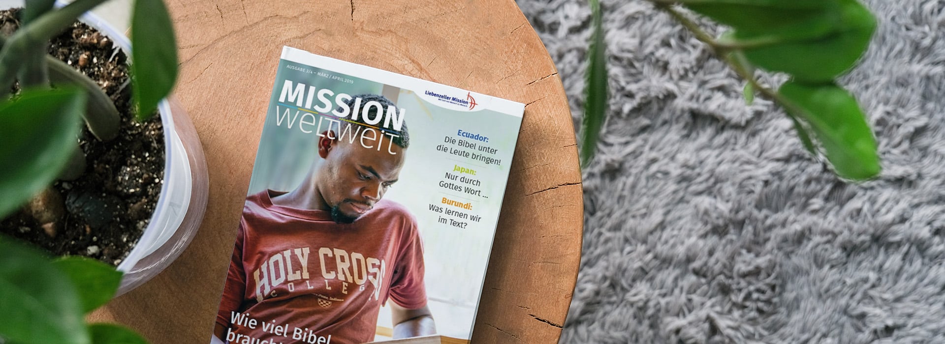 Auf einem Holztisch neben grünen Pflanzen und einem grauen Teppich liegt eine deutsche Zeitschrift mit dem Titel „Mission weltweit“.