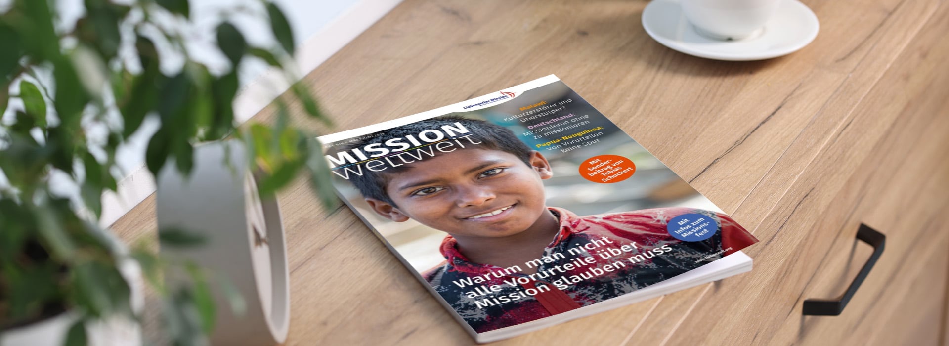 Eine Zeitschrift mit dem Titel „Mission Weltweit“, auf deren Titelseite ein lächelnder Junge zu sehen ist, liegt auf einem Holztisch neben einer Pflanze und einer weißen Tasse.