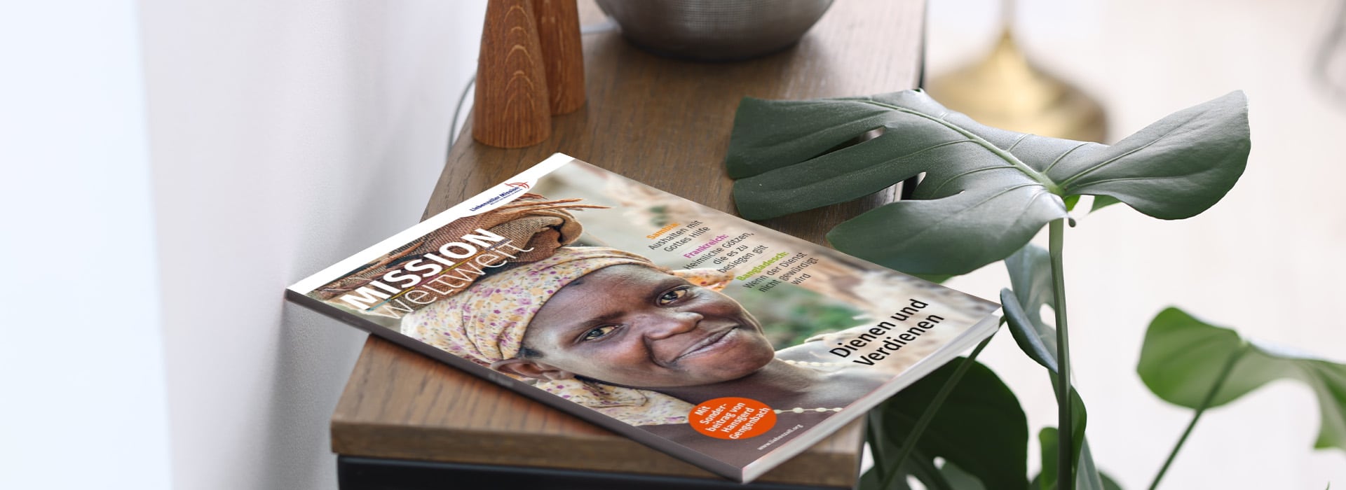 Die Zeitschrift „Mission Weltweit“ mit einer lächelnden Frau auf dem Titelblatt liegt auf einem Holztisch neben einer grünen Pflanze.