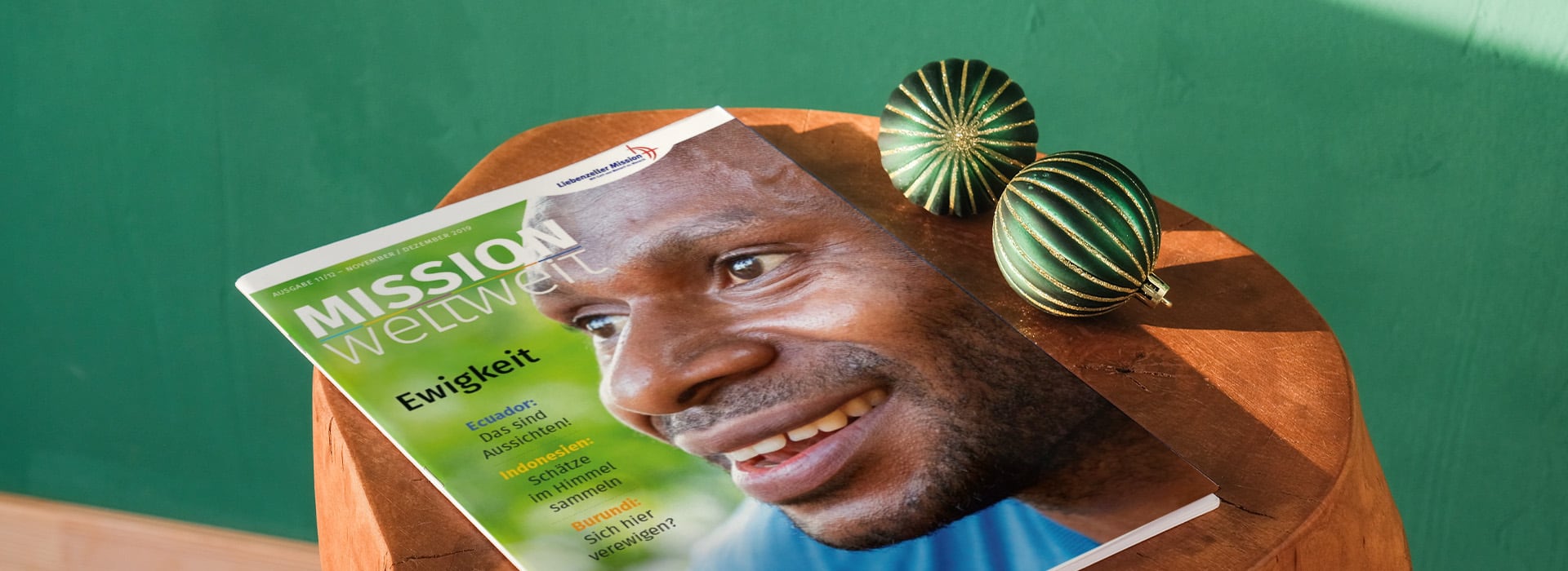 Eine Zeitschrift mit dem Titel „MISSION Weltweit“, auf deren Titelseite ein lächelnder Mann zu sehen ist, liegt auf einem Holztisch neben zwei grünen Dekoartikeln.