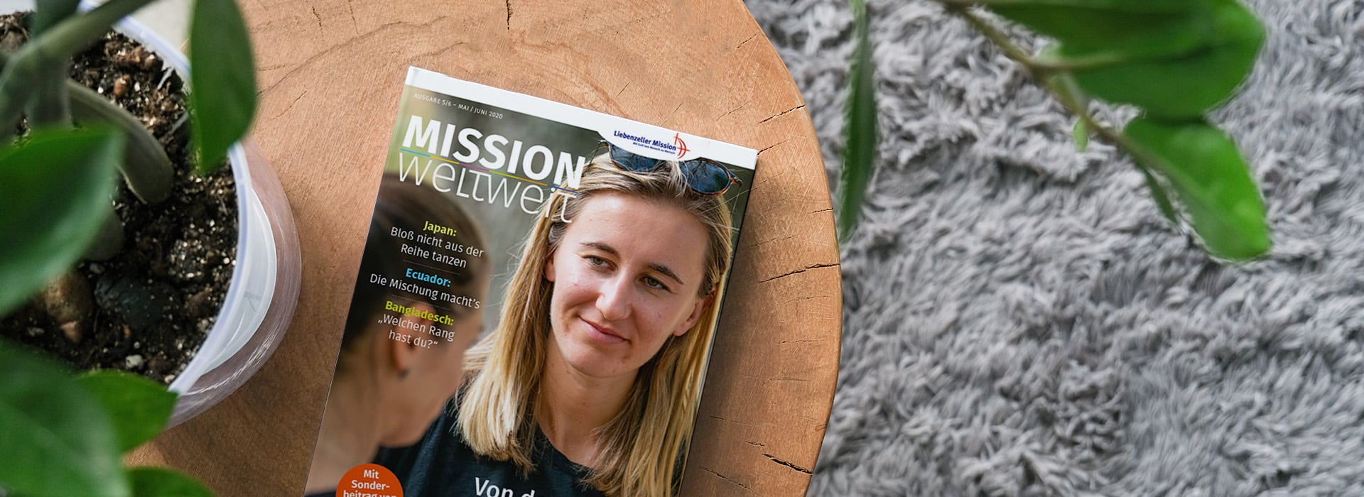 Auf einem Holztisch neben einer Topfpflanze und einem grauen Teppich liegt eine Zeitschrift mit dem Foto einer Frau auf dem Cover.