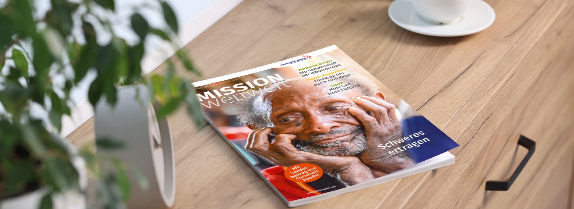 Auf einem Holztisch neben einer Pflanze liegt eine Zeitschrift mit dem Titel „MISSION weltweit“, auf deren Titelseite ein nachdenklicher älterer Mensch abgebildet ist.