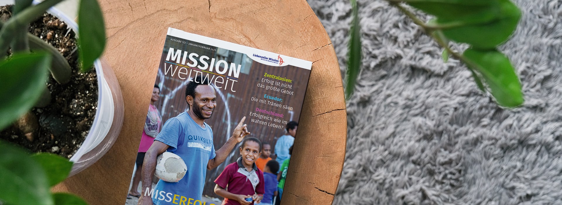 Die Zeitschrift „Mission weltweit“ liegt auf einem Holztisch, neben einer Topfpflanze und einem grauen Teppich.