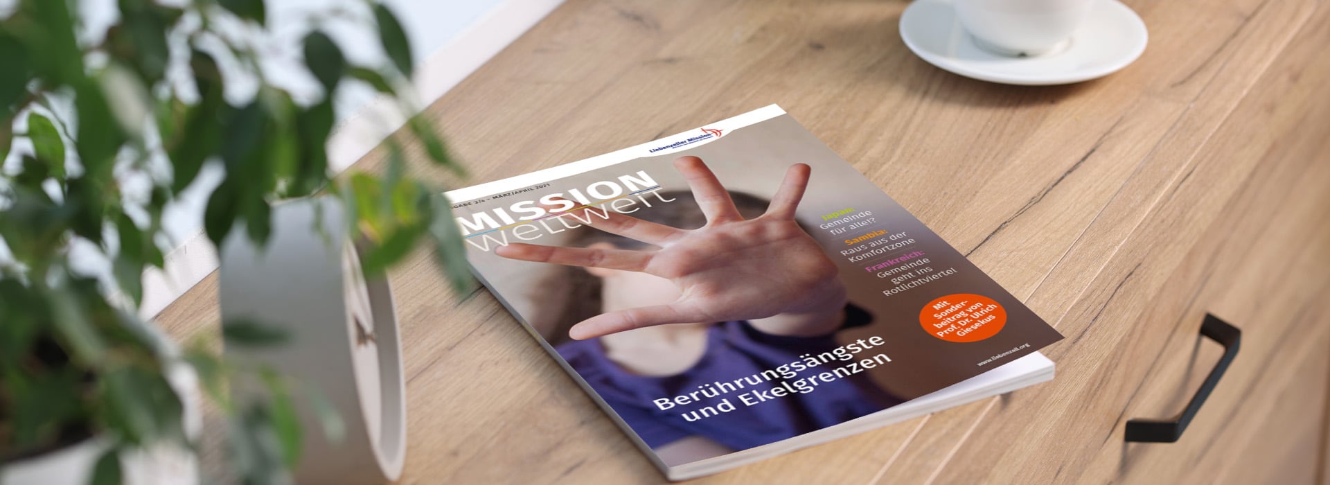 Die Zeitschrift „Mission weltweit“ liegt auf einem Holztisch, neben einer Pflanze und einer weißen Kaffeetasse mit Untertasse.