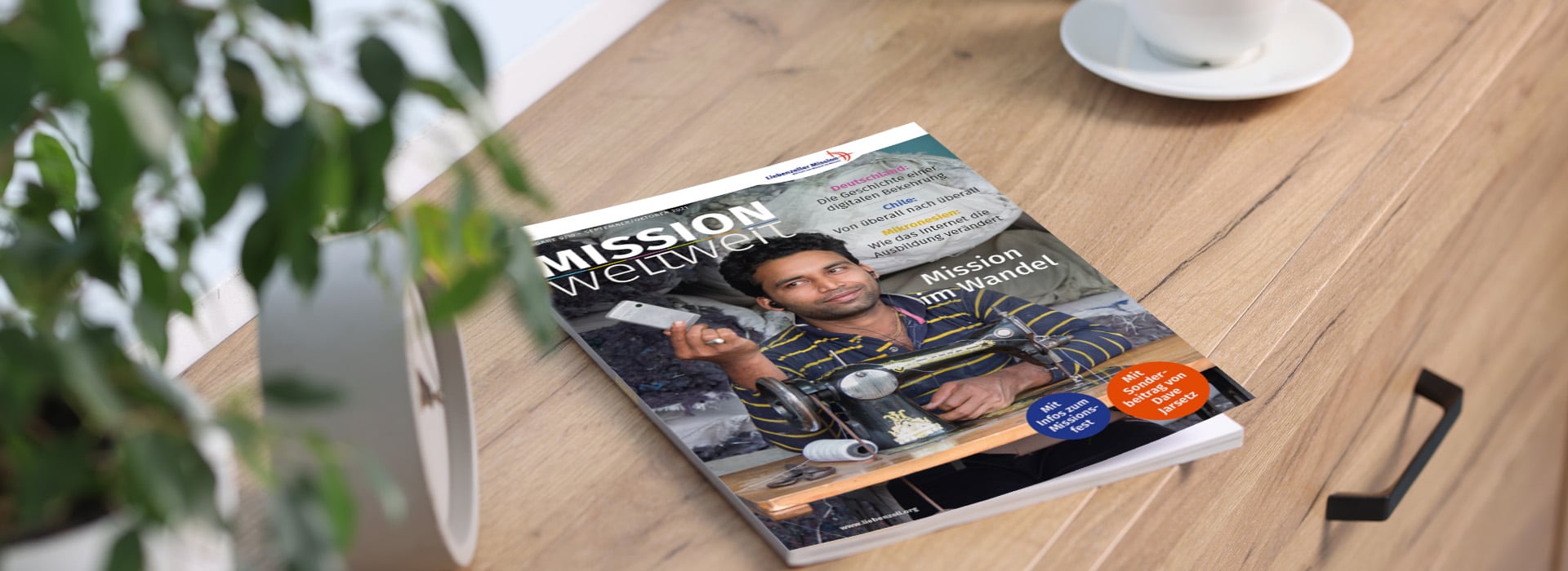 Auf einem Holztisch neben einer Pflanze und einer weißen Tasse liegt eine Zeitschrift mit dem Titel „MISSION Weltweit“.