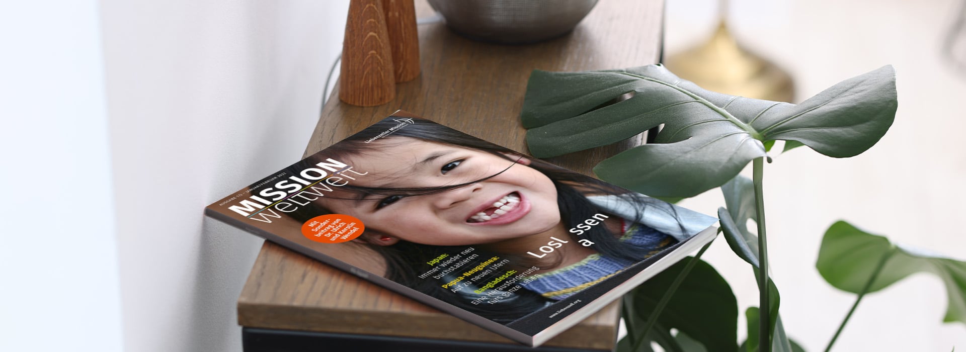 Eine Zeitschrift mit einem lächelnden Kind auf dem Cover, die auf einer Holzunterlage neben einer grünen Pflanze und einer dekorativen Vase liegt.