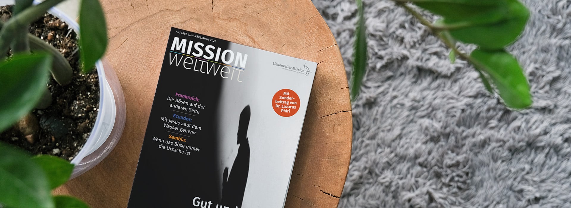 Auf einem Holztisch neben einer Topfpflanze und einem grauen Teppich liegt eine deutsche Zeitschrift mit dem Titel „Mission Weltweit“.