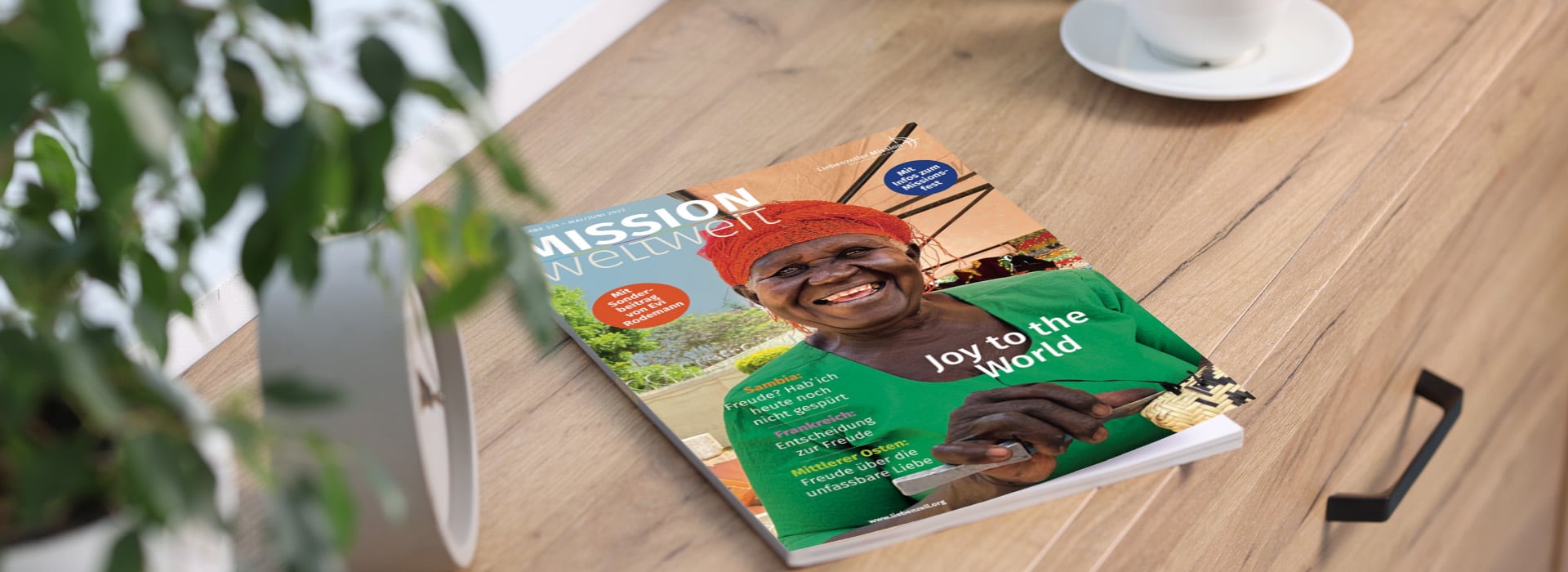 Eine Zeitschrift mit dem Titel „Mission Weltweit“, auf deren Titelseite eine lächelnde Person mit einem roten Kopftuch zu sehen ist, die auf einem Holztisch liegt.