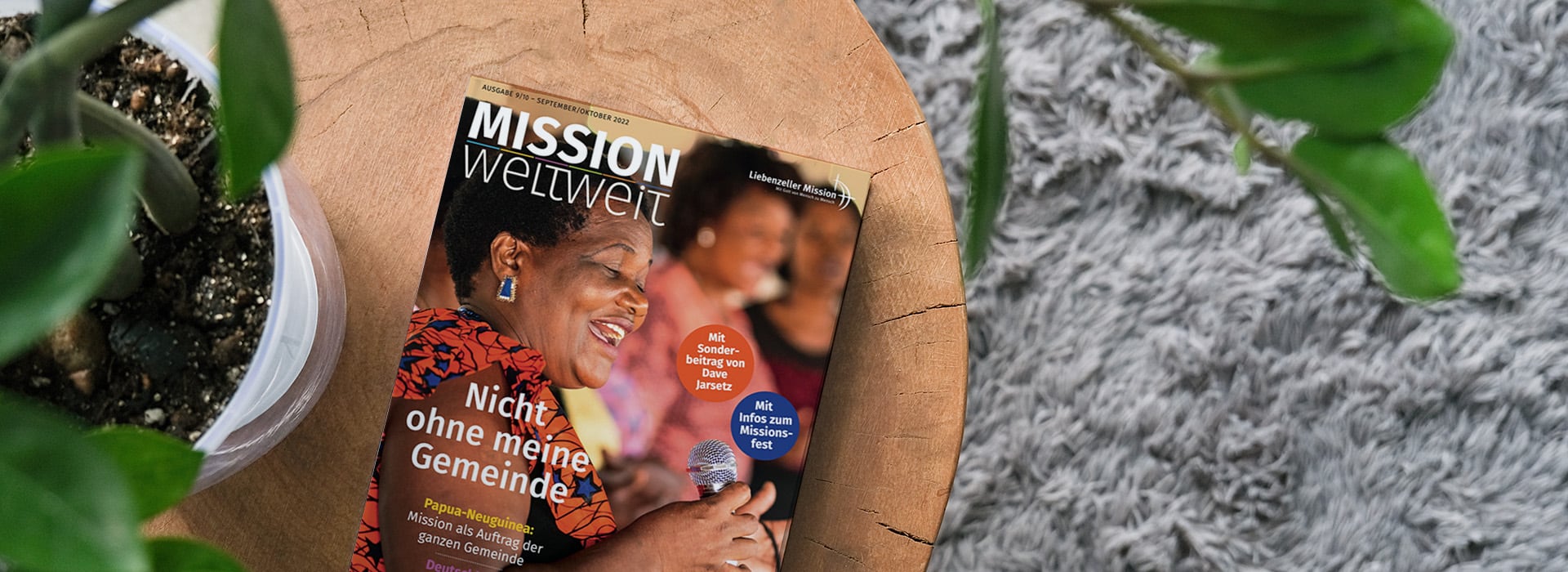 Auf einem Holztisch neben einer Topfpflanze und einem grauen Teppich liegt eine Zeitschrift mit dem Titel „MISSION weltweit“.