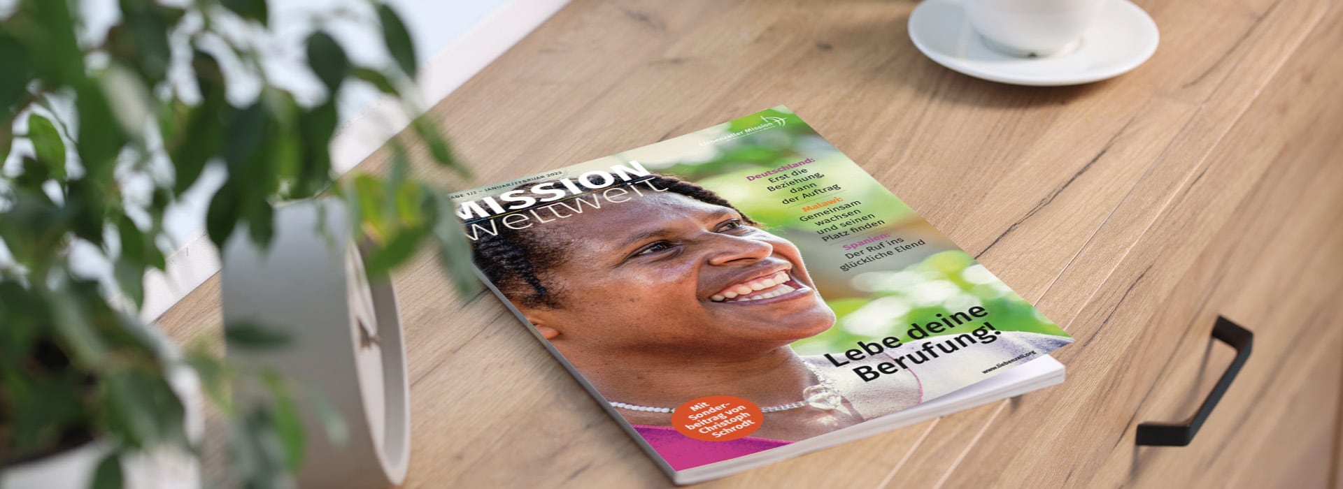 Eine Zeitschrift mit dem Titel „Mission Weltweit“, auf deren Titelseite eine lächelnde Frau zu sehen ist, liegt auf einem Holztisch neben einer Pflanze und einer Kaffeetasse.