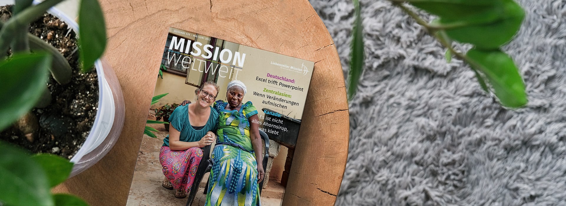 Eine Zeitschrift mit dem Titel „MISSION weltweit“ auf einem Holztisch neben einer Pflanze; auf dem Titelblatt sind zwei lächelnde Frauen zu sehen.