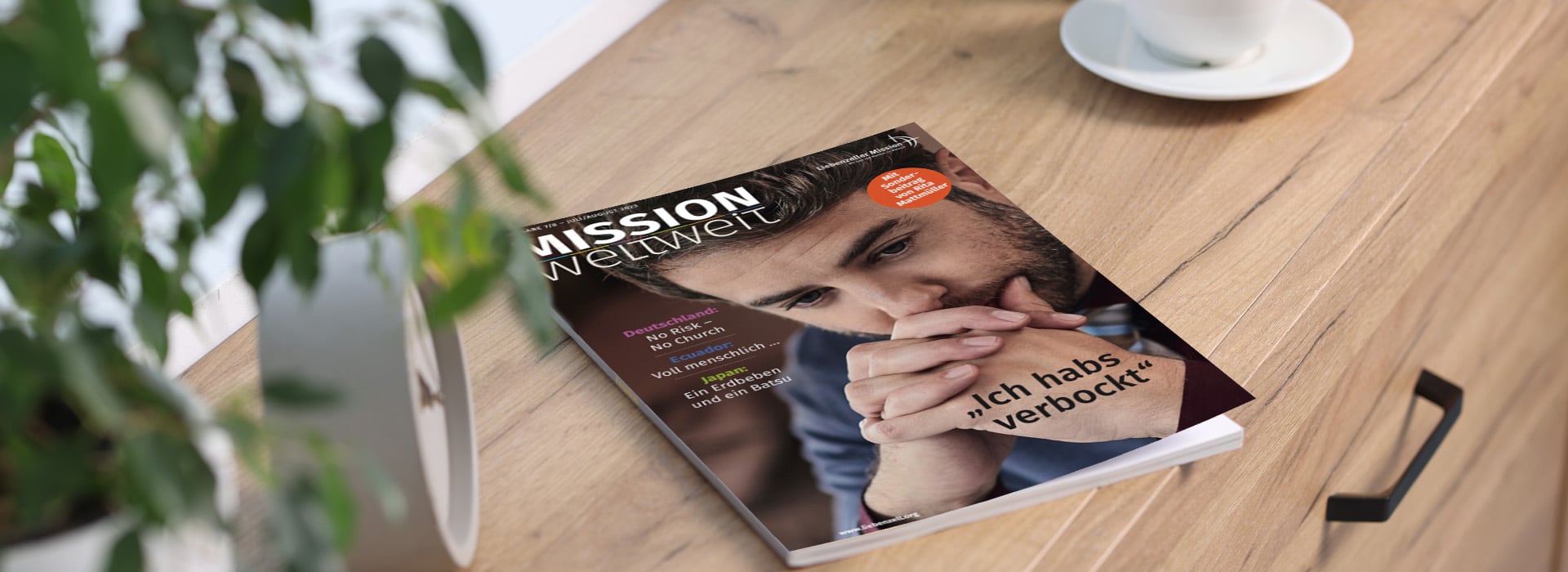 Auf einem Holztisch liegen neben einer Pflanze und einer weißen Tasse eine deutsche Zeitschrift mit dem Titel „Mission Weltweit“.