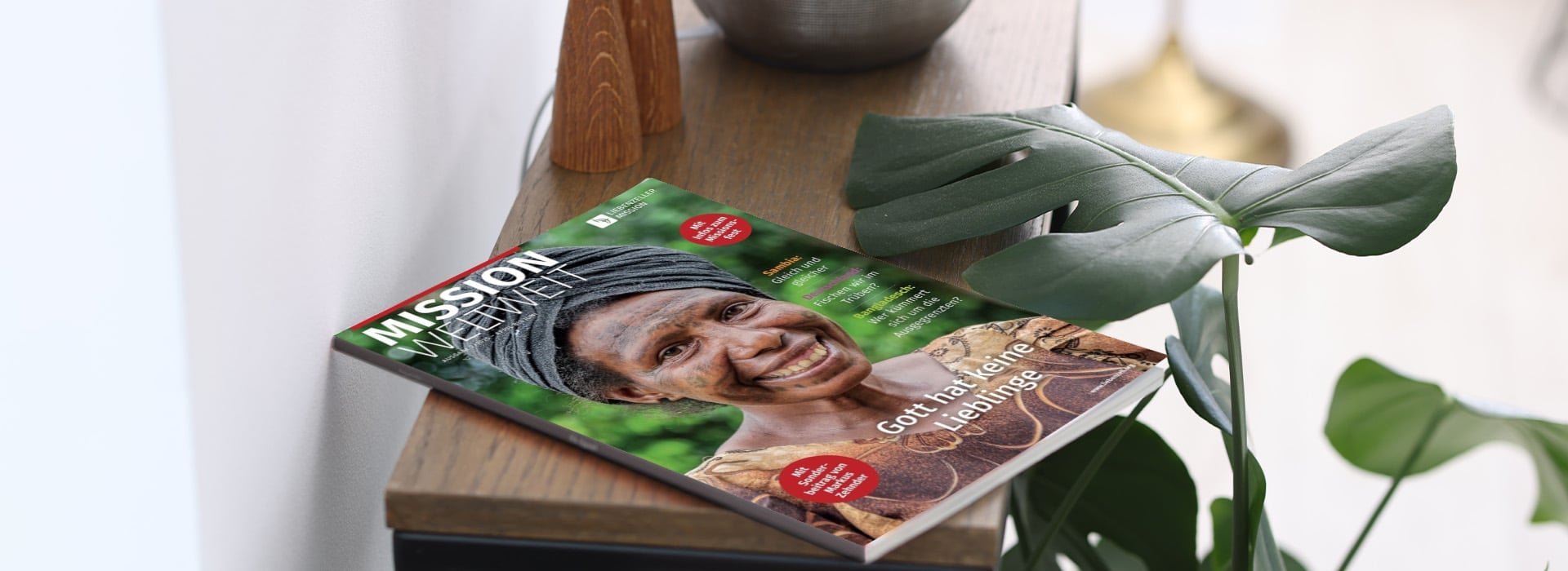 Eine Zeitschrift mit dem Titel „MISSION weltweit“, auf deren Titelseite eine lächelnde Frau zu sehen ist, liegt auf einem Holztisch neben einer grünen Pflanze.