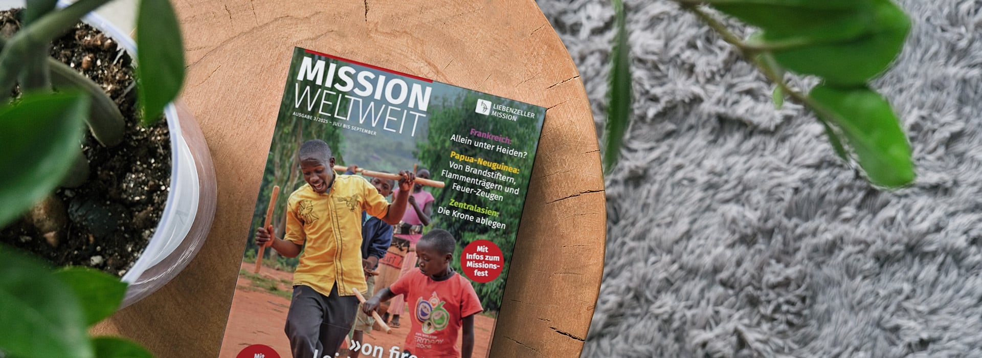 Auf einem Holztisch neben einer Topfpflanze und einem grauen Teppich liegt eine Zeitschrift mit dem Titel „Mission Weltweit“.