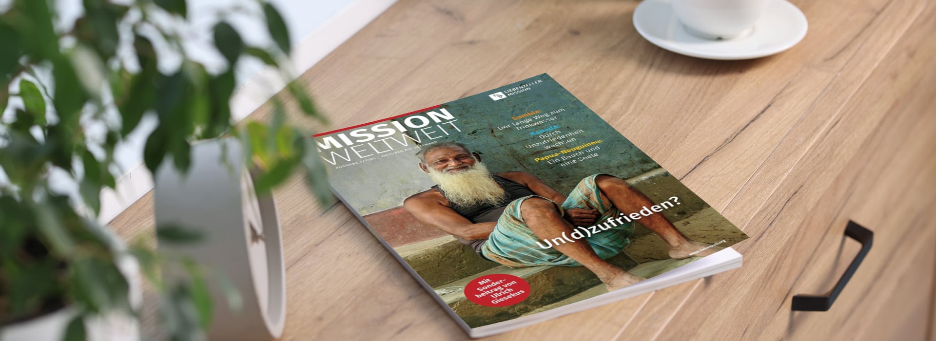 Auf einem Holztisch neben einer Pflanze und einer Kaffeetasse liegt eine Zeitschrift mit dem Titel „MISSION WELTWEIT“, auf deren Titelseite ein älterer Mann abgebildet ist.