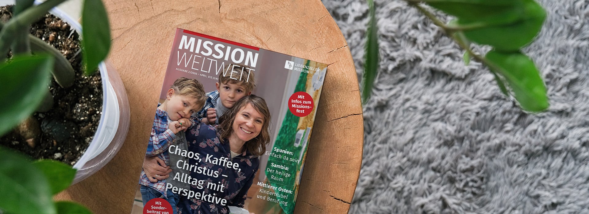 Auf einem Holztisch neben einer Topfpflanze und einem grauen Teppich liegt eine Zeitschrift mit dem Titel „MISSION WELTWEIT“.