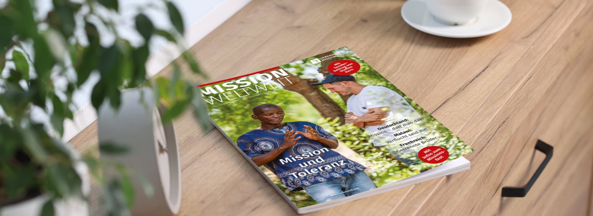 Auf einem Holztisch neben einer Topfpflanze und einer weißen Tasse liegt eine Zeitschrift, auf deren Titelseite zwei Personen zu sehen sind.