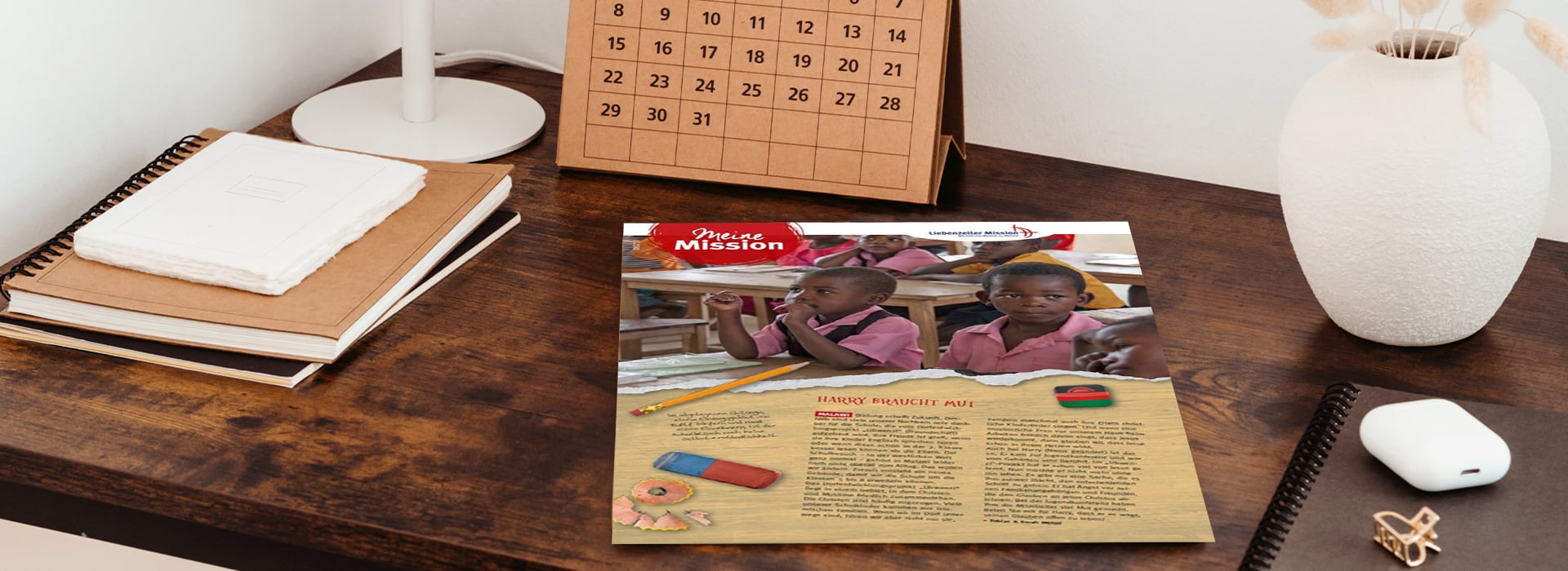Auf einem Holztisch liegen ein Flyer mit Kindern in einem Klassenzimmer sowie Hefte, ein Kalender und eine weiße Vase.