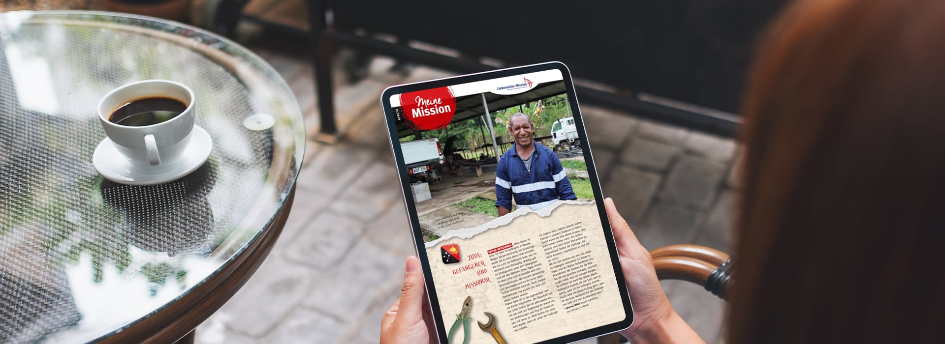 Eine Person hält ein Tablet in der Hand, auf dem ein Artikel mit dem Foto eines lächelnden Mannes zu sehen ist, daneben steht ein Tisch mit einer Tasse Kaffee.