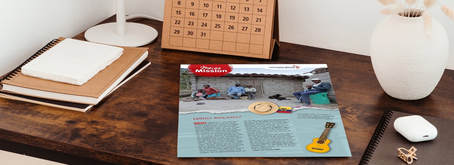 Auf einem Holztisch liegt neben Notizbüchern, einem Kalender und einer Pflanze eine Zeitschrift mit dem Foto von vier Personen, die im Freien sitzen.