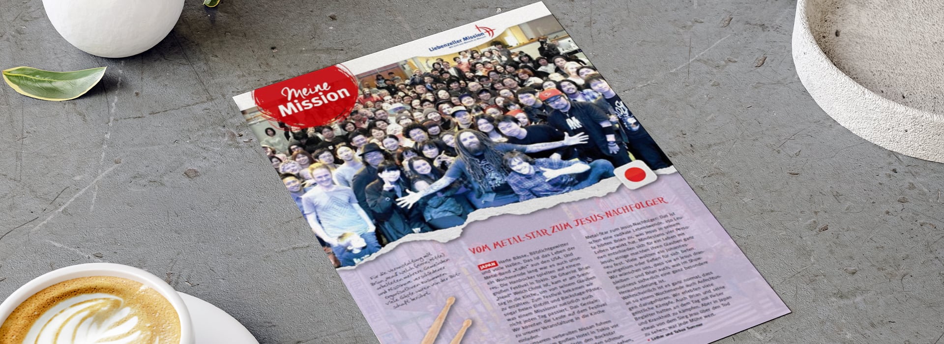 Auf einer Zeitschriftenseite, die auf einem grauen Tisch liegt, sind ein großes Gruppenfoto und ein deutscher Text zu sehen, daneben stehen eine Kaffeetasse und eine kleine Pflanze.