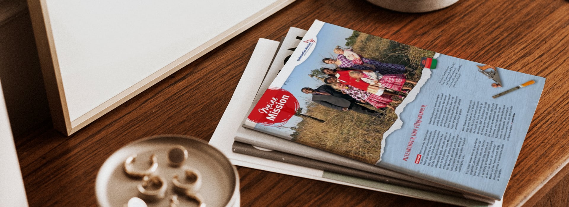 Auf einem Holztisch liegt neben einer Tasse und anderen Papieren eine Broschüre mit einem Familienfoto und dem Text „Unsere Mission“.