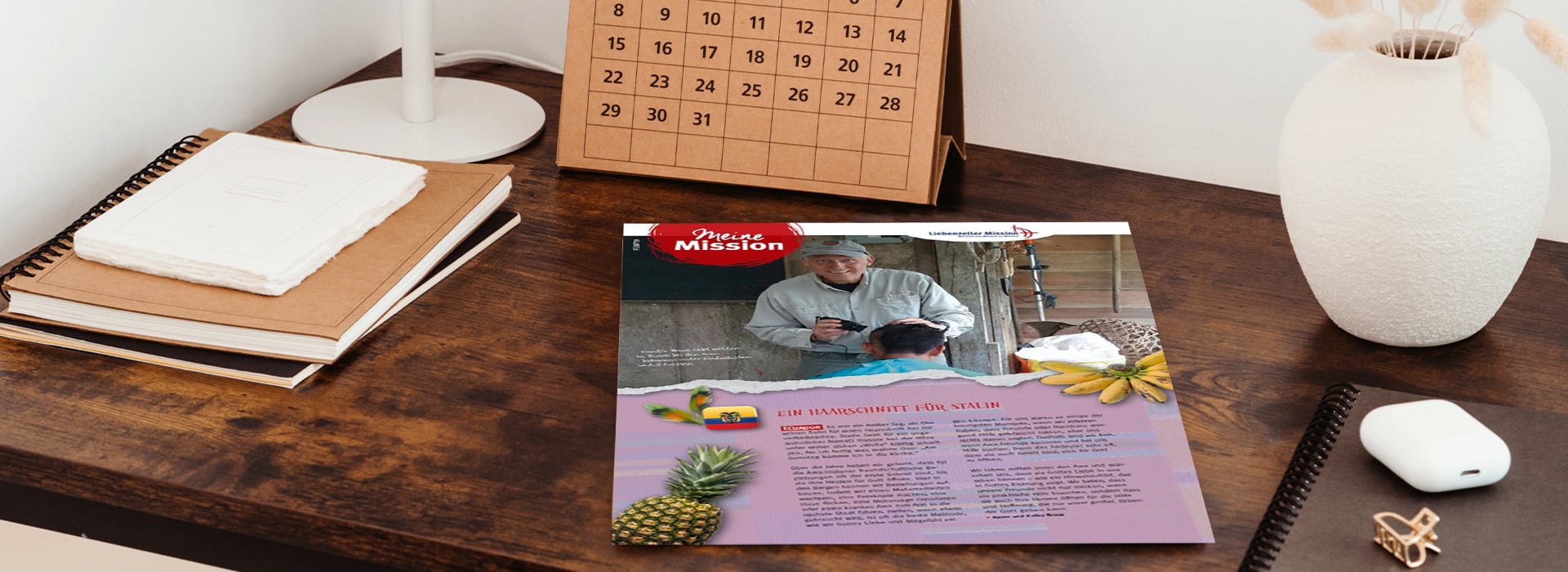 Auf einem Schreibtisch, auf dem Notizbücher, ein Kalender und eine Vase stehen, liegt ein Newsletter, auf dem zwei Personen, Ananas und Text zu sehen sind.