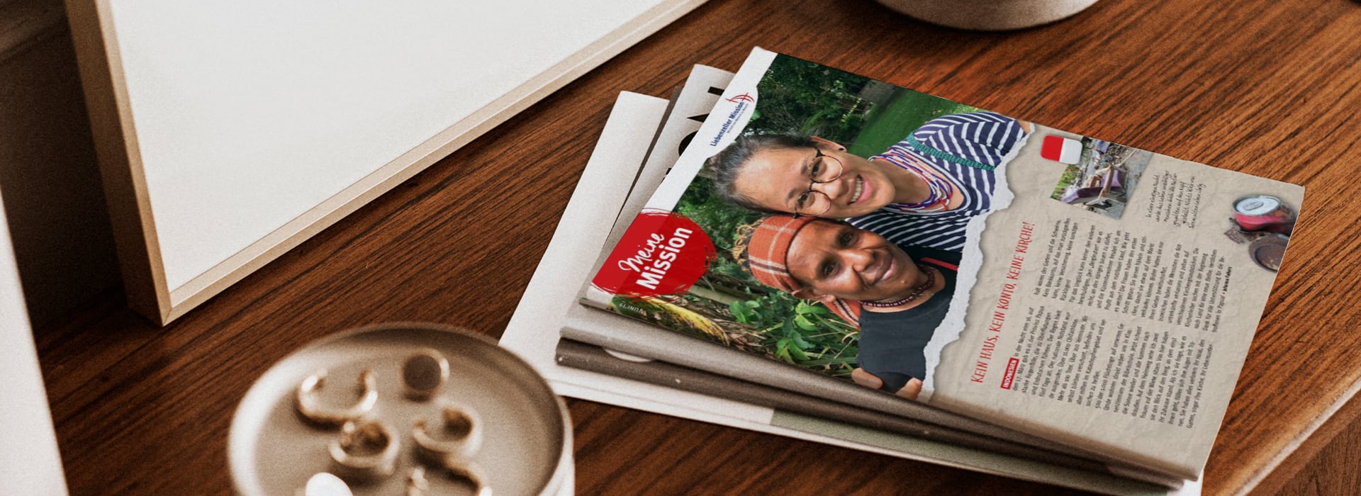 Auf einem Holztisch liegt eine Zeitschrift mit zwei lächelnden Frauen auf dem Cover, daneben stehen ein Getränk und ein weißer Gegenstand.