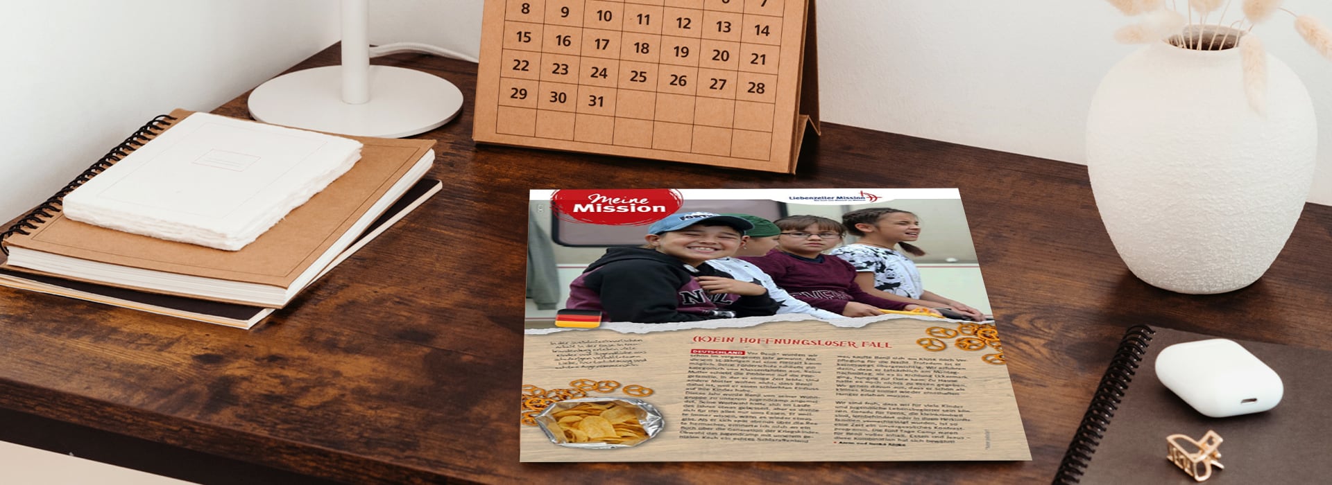 Ein Flyer mit drei lächelnden Kindern auf einem Schreibtisch neben einem Kalender, einem Notizblock und einer Lampe.